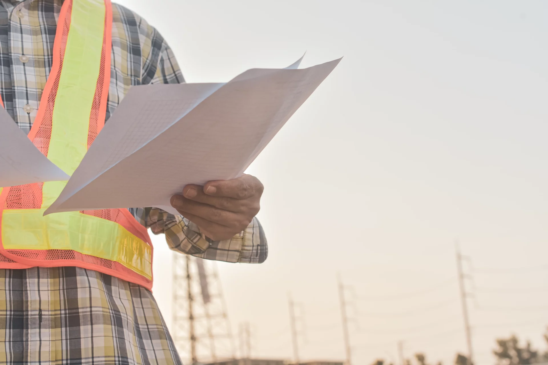 A man in constrution cloting standing on a work site holding a document.